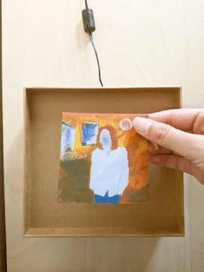 A wooden shadow box on a wall, with a hand holding a painted RFID card