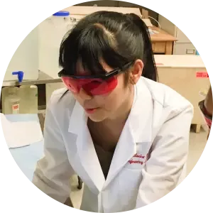 Nhi in a lab coat and red safety glasses, working at a bench